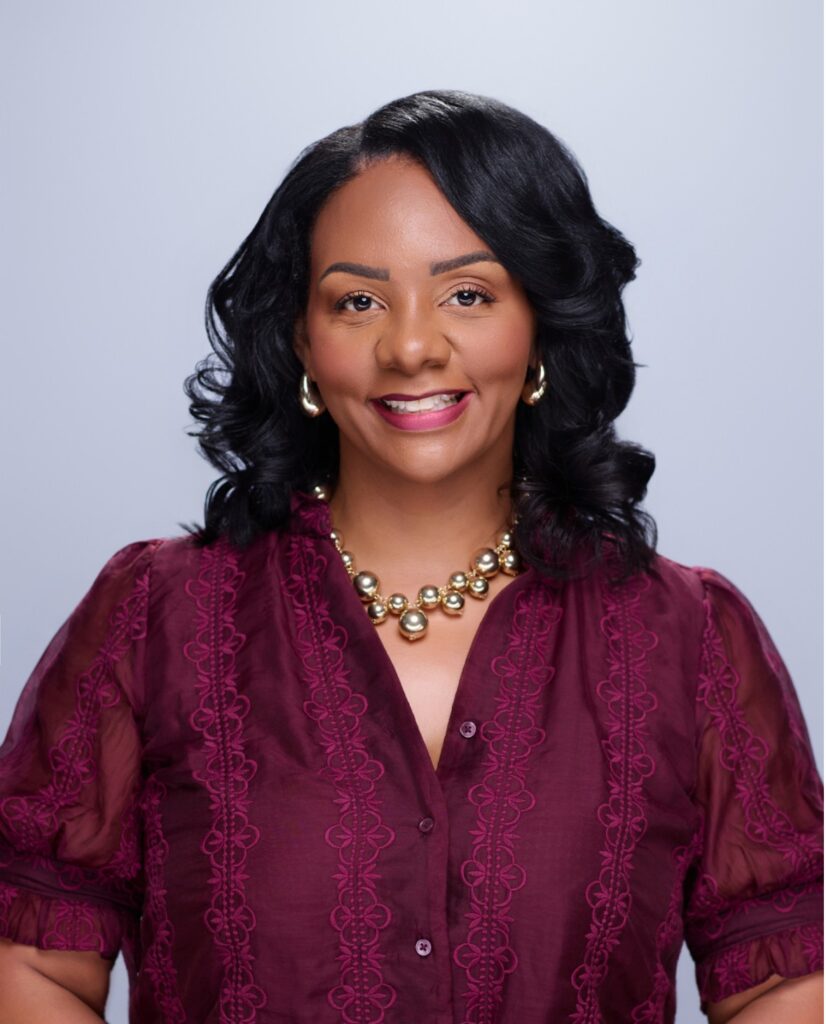 Smiling woman with black hair in a maroon embroidered blouse and gold jewelry, posed against a light gray background.